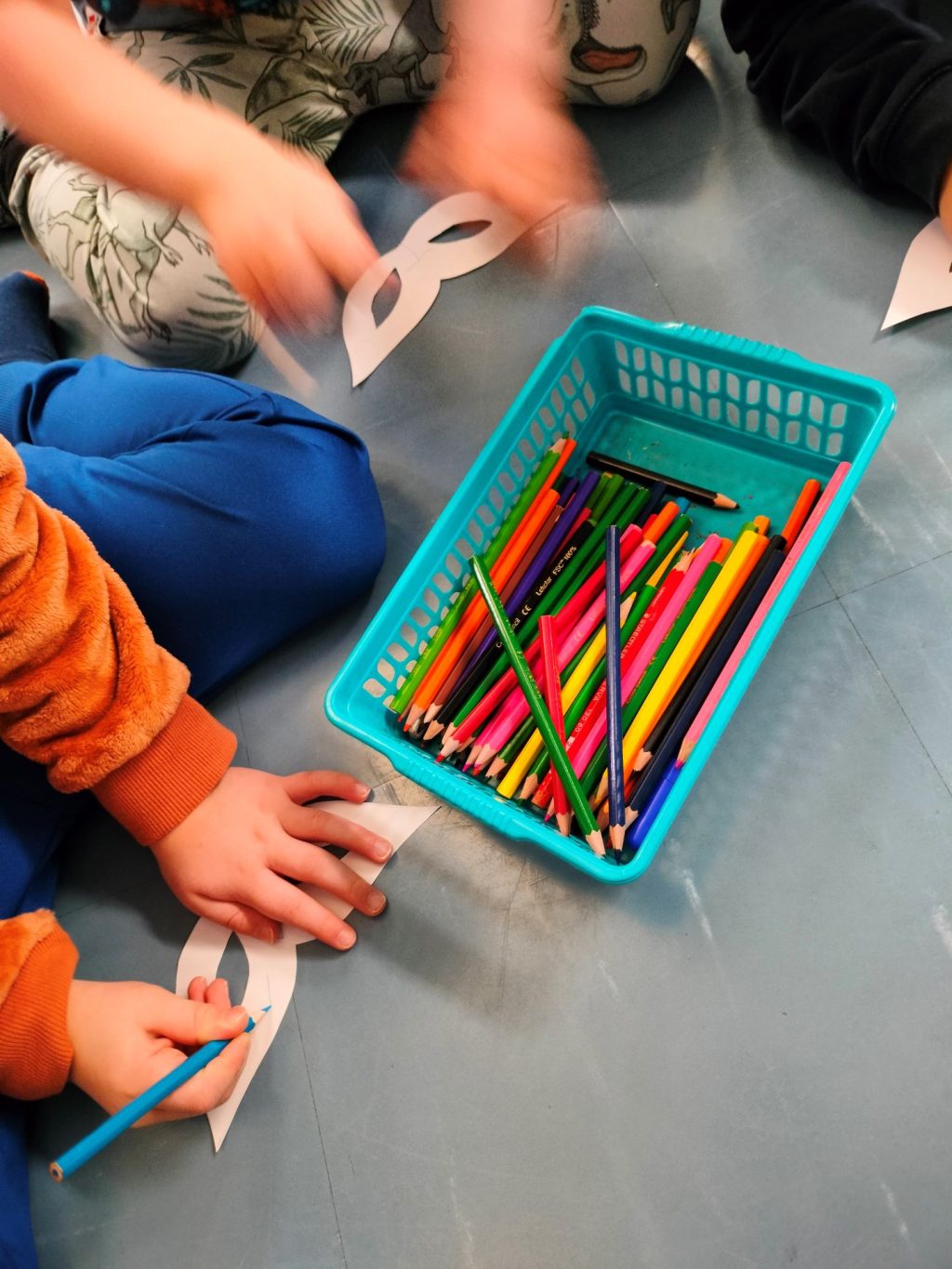 Keskellä värikynälaatikko. Reunassa kahden lapsen kädet värittämässä paperista leikattua maskia.
