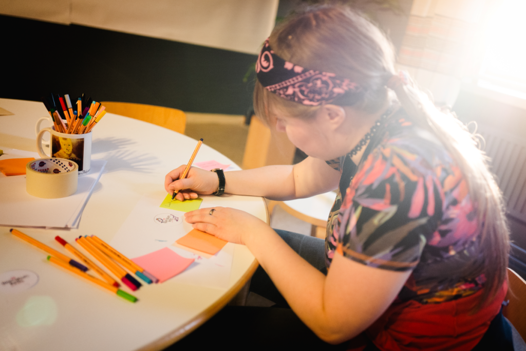 Tyttö/nuori nainen piirtää post it -lappuun pöydän ääressä. Pöydällä värikyniä, teippirulla ja paperia.