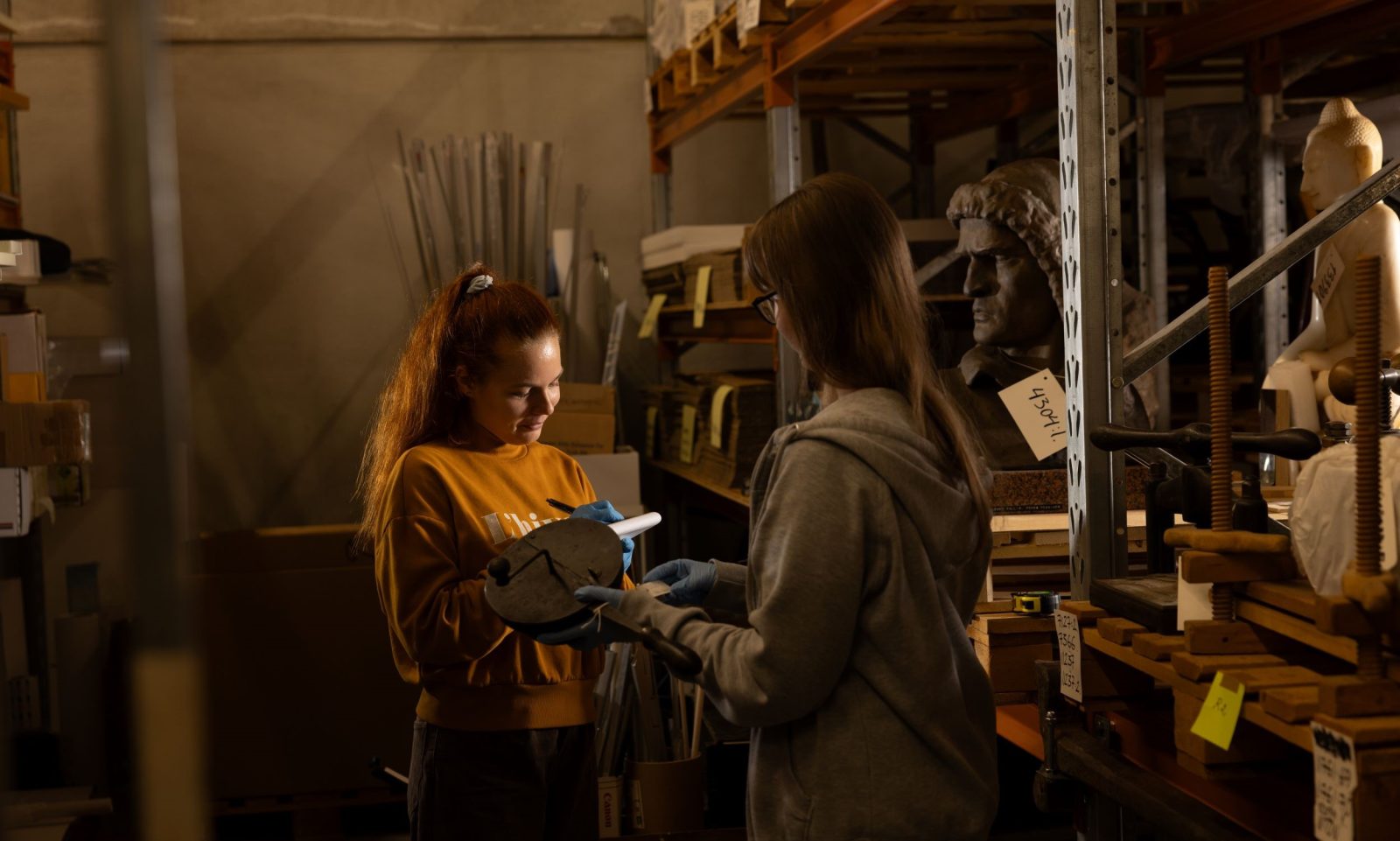 Kaksi naista; toinen kirjoittaa lehtiöön ja toisella museoesine käsissä. Taustalla varastohyllyissä tavaroita, joissa on kiinni numerolappuja.