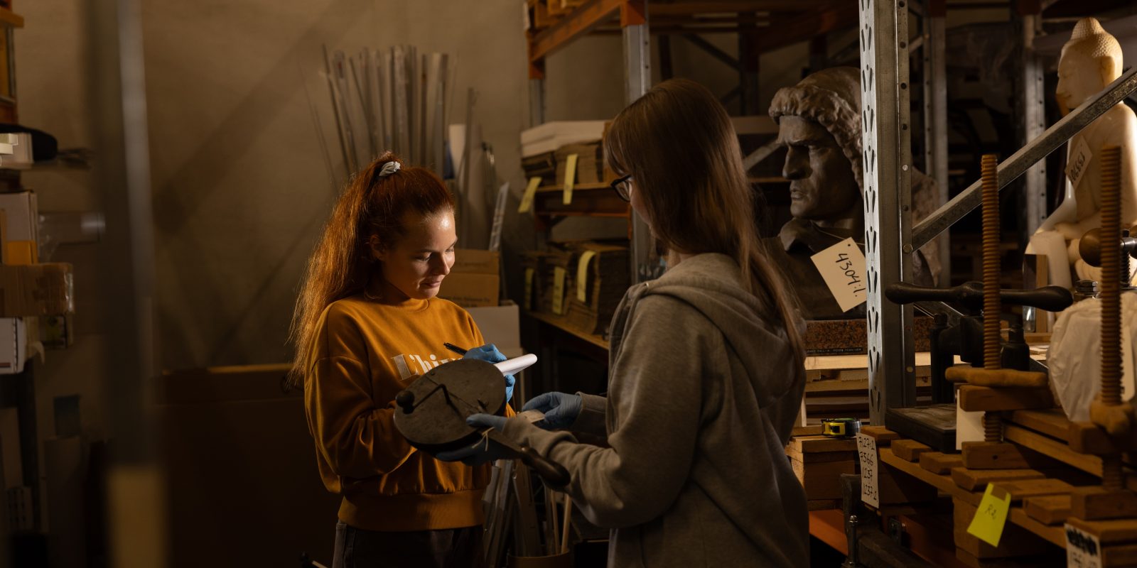 Two women, one in red hair and orange sweatshirt, the other in grey swater and brown hair. They are in a warehouse, two statues in the shelf. One woman is writing down somethin as the other one is holding something in her hands.