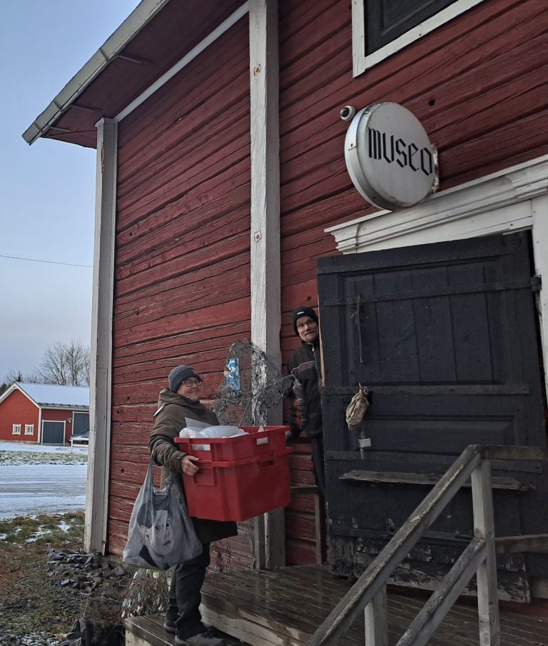 Punainen vanha rakennus, musta ovi, jonka päällä kyltti "museo". Ovesta kurkistaa mies. Portailla seisoo nainen iso punainen laatikko käsissään.