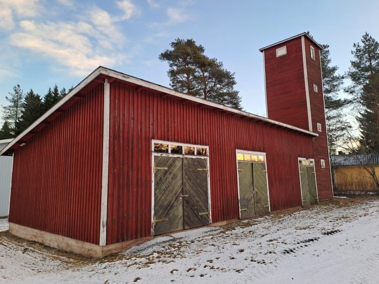 Punainen, puinen ja pitkänmuotoinen rakennus. Pitkällä sivulla kolme kaksoisovea ja toisessa päässä tornimainen osa.