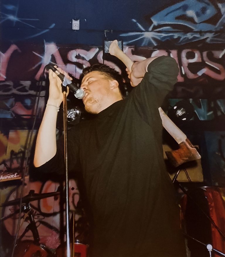 Mikko Laurila is singing while holding an axe behind his back. In the background, the venue's dark wall is decorated with graffiti.