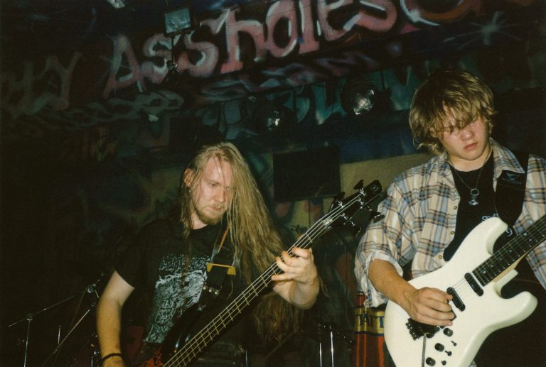 Guitarist Miika Tenkula is playing on the right side of the frame, while bassist Niko Karppinen is positioned in the center. In the background, the venue's dark, graffiti-covered surroundings are visible.